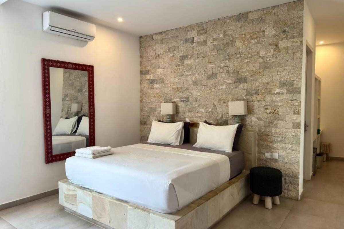Ein Schlafzimmer mit einer Steinwand, einem Bett mit weißer Bettwäsche, einem Spiegel mit rotem Rahmen und einem kleinen Hocker.