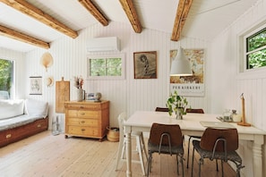 Ein Zimmer mit einem weißen Holztisch, vier Stühlen, einer Holzkommode, einer Bank, einem Fenster und einem an der Wand hängenden Bild.