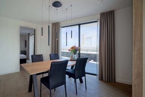 A modern dining area with a wooden table and black chairs, large windows with curtains, and a view of the cityscape. - Corner Apartment With Harbor Views (Breskens)