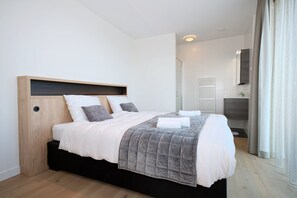 A modern hotel room with a bed, a built-in shelf, a small bedside table, and a bathroom with a shower and toilet. - Corner Apartment With Harbor Views (Breskens)