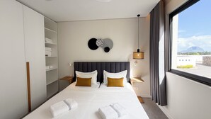 A modern hotel room with a bed, a ceiling fan, a wall-mounted light, a window with a view, and a wardrobe. - Asmara Residence- 2 Bedroom,Grande Riviere Noire (Grande Riviere Noire)