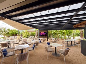 Un patio exterior con varias mesas y sillas, una pantalla de televisión y vista al océano.