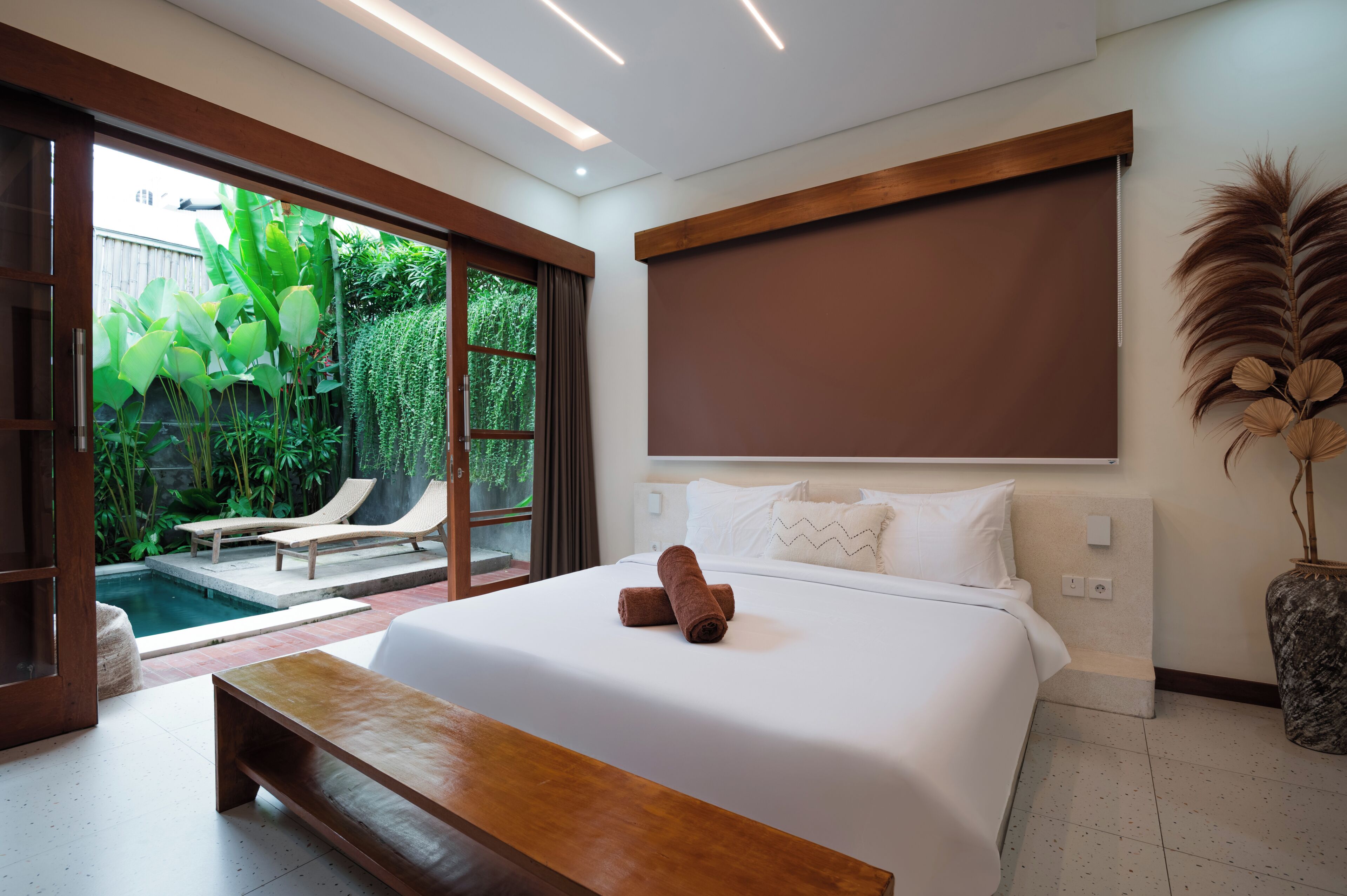 Habitación de hotel moderna con una cama grande, un banco, vista a una piscina y un ventanal corredizo que da a un jardín.