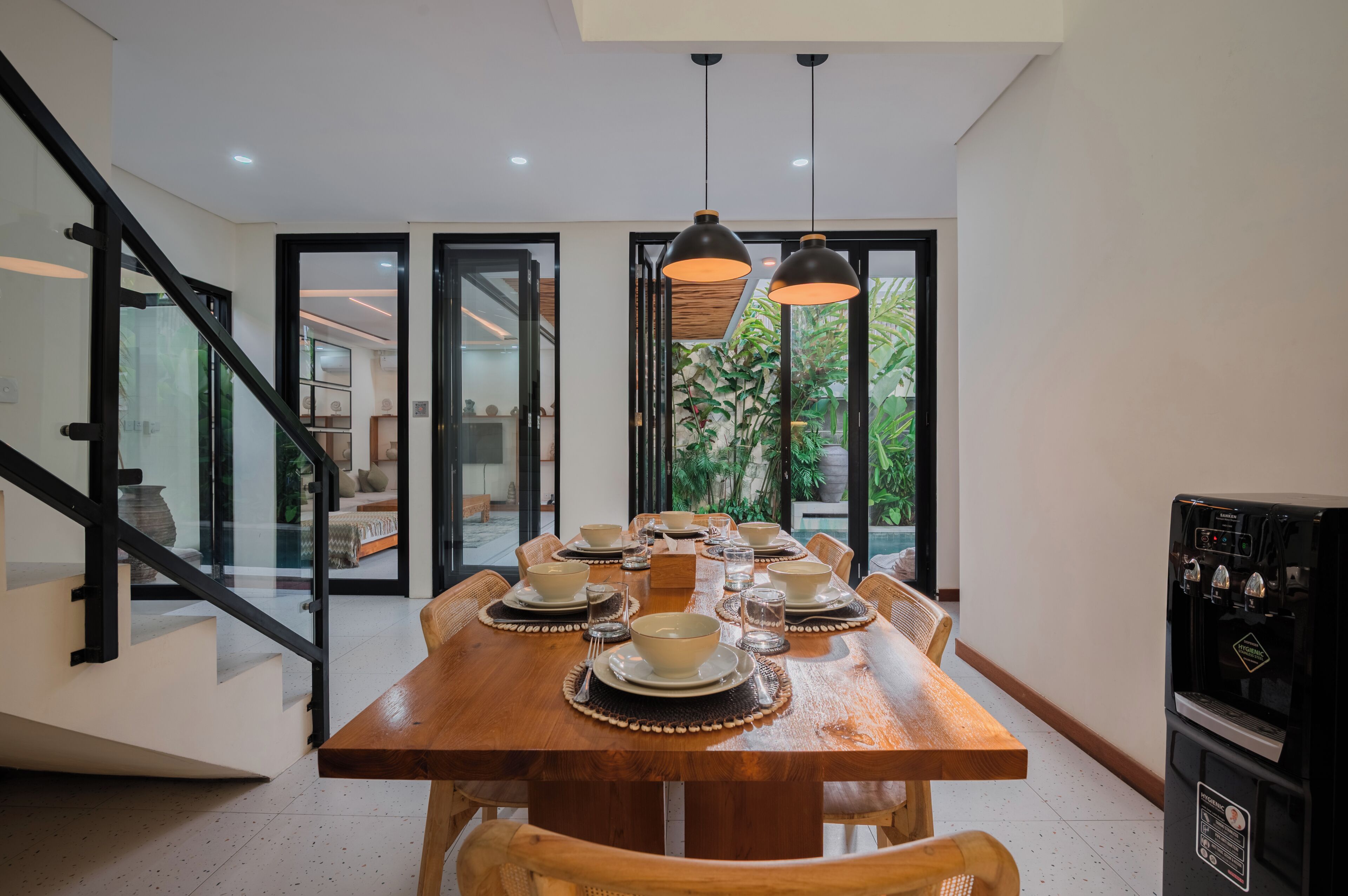 A dining area with a wooden table set for six, a water dispenser, and a staircase with glass doors.