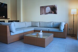 Un sofá modular con cojines, una mesa de centro con cubierta de vidrio, un televisor montado en la pared y una lámpara de pie.