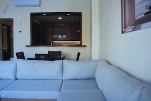 Un salon moderne avec un canapĂ© dâangle, un coin repas avec des chaises, une cuisine avec une fenĂȘtre et un climatiseur fixĂ© au mur.