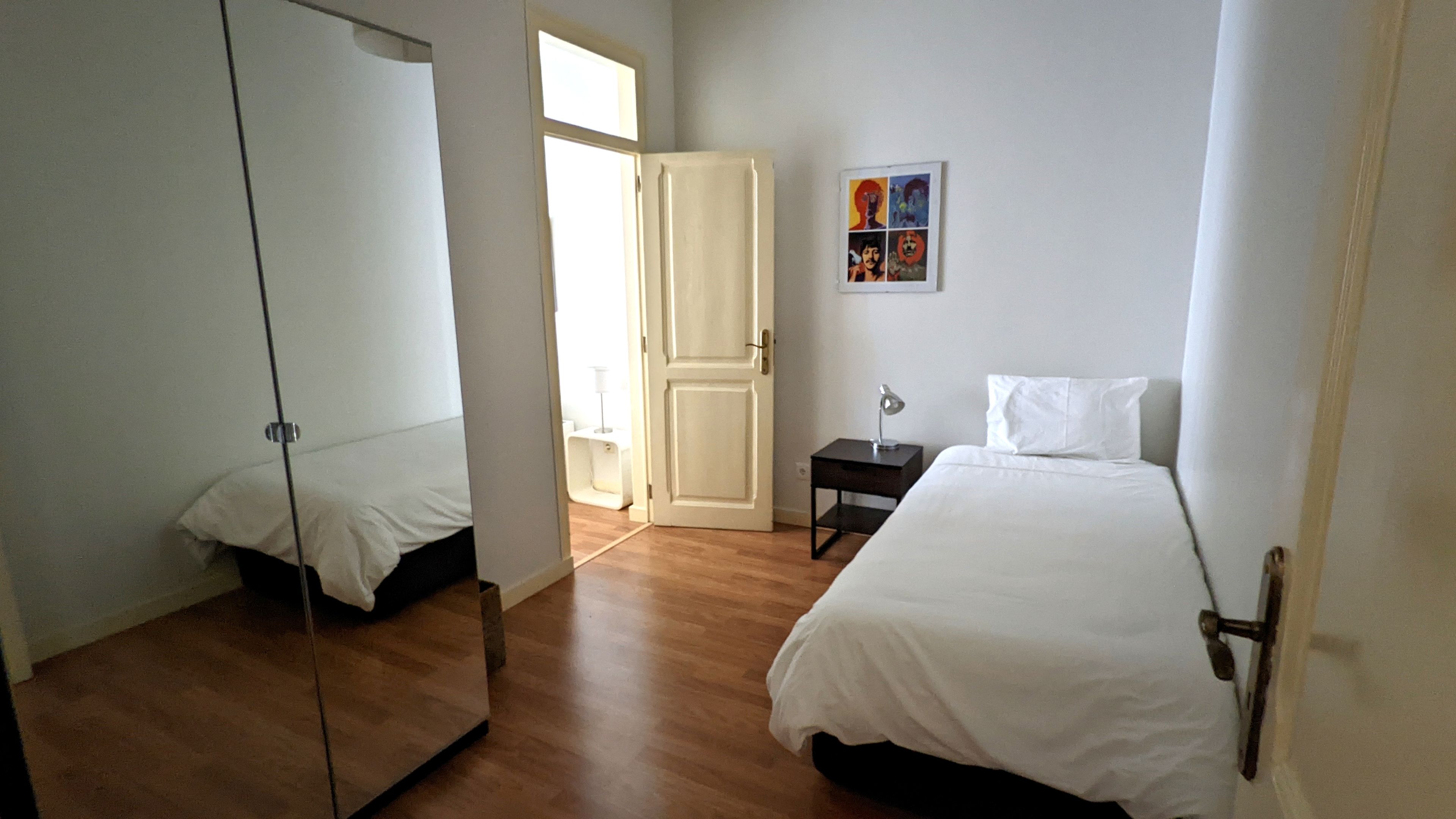 A bedroom with a bed, a bedside table, a lamp, a mirror, and a door leading to another room.