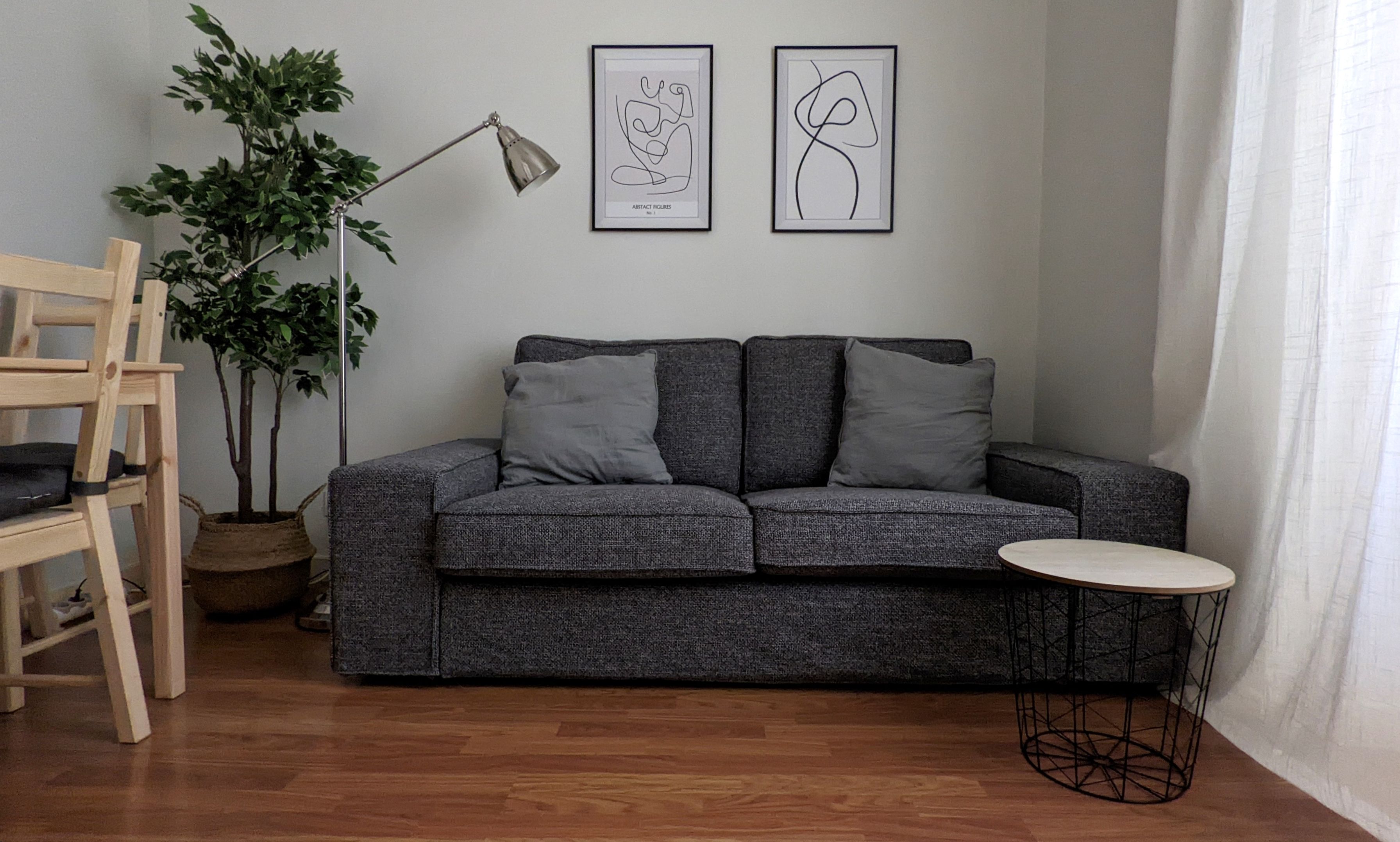 Een moderne woonkamer met een grijze bank, twee kussens, een kleine ronde tafel, een vloerlamp en twee abstracte kunstwerken aan de muur.