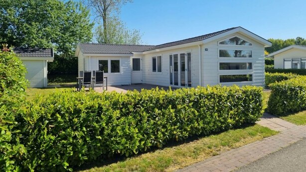 A single-story mobile home with a garage, a patio area with chairs, and a well-maintained hedge in front. - Holiday Home in Velsen-zuid With Terrace (Velsen-Zuid)