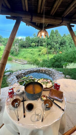 Une table à manger, ornée d’un pot de soupe, de différents plats et de condiments, se dressant sur fond d’un paysage verdoyant luxuriant et d’un ciel dégagé.