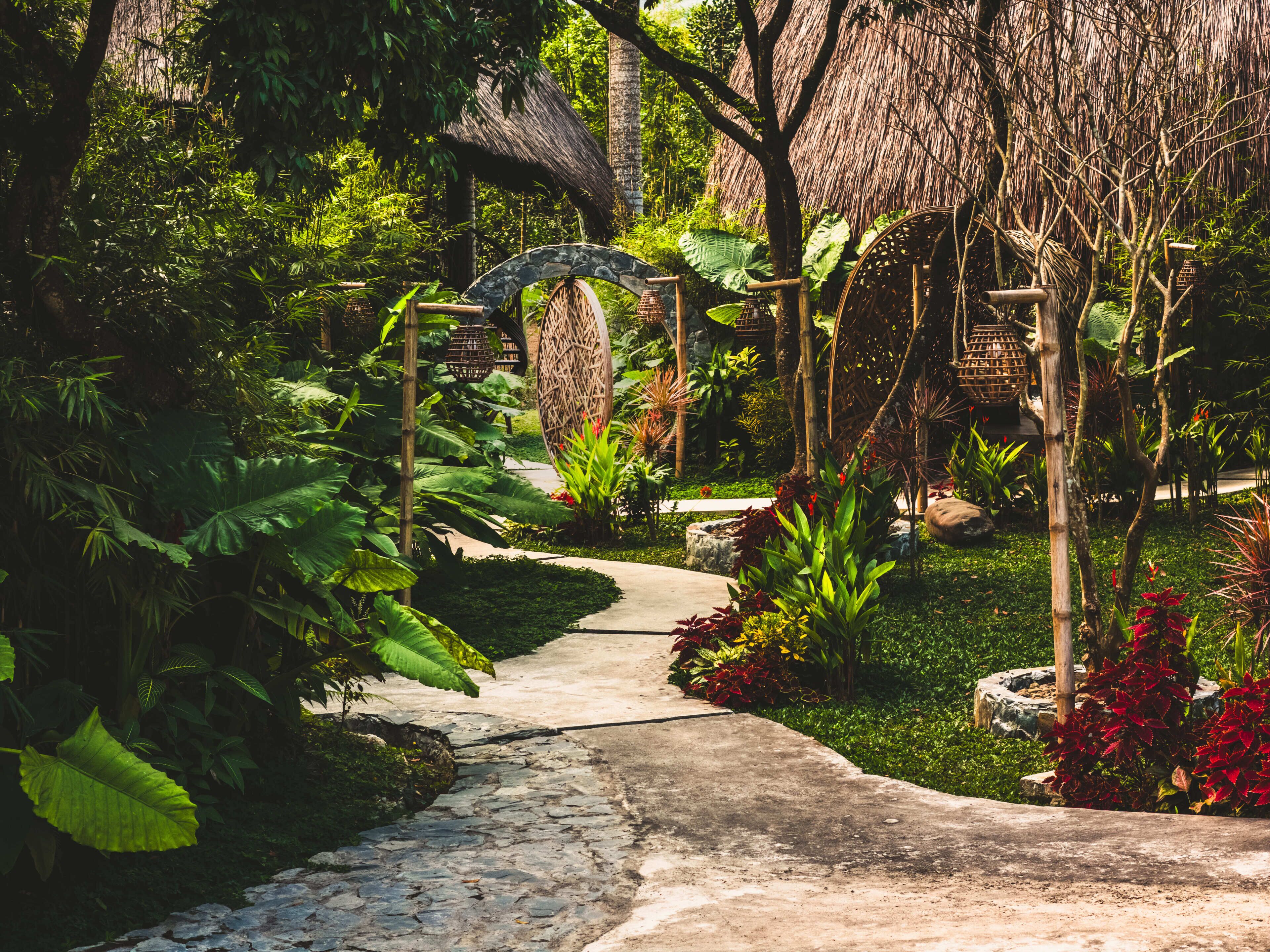 A garden pathway with a thatched roof structure, stone paving, and tropical vegetation.