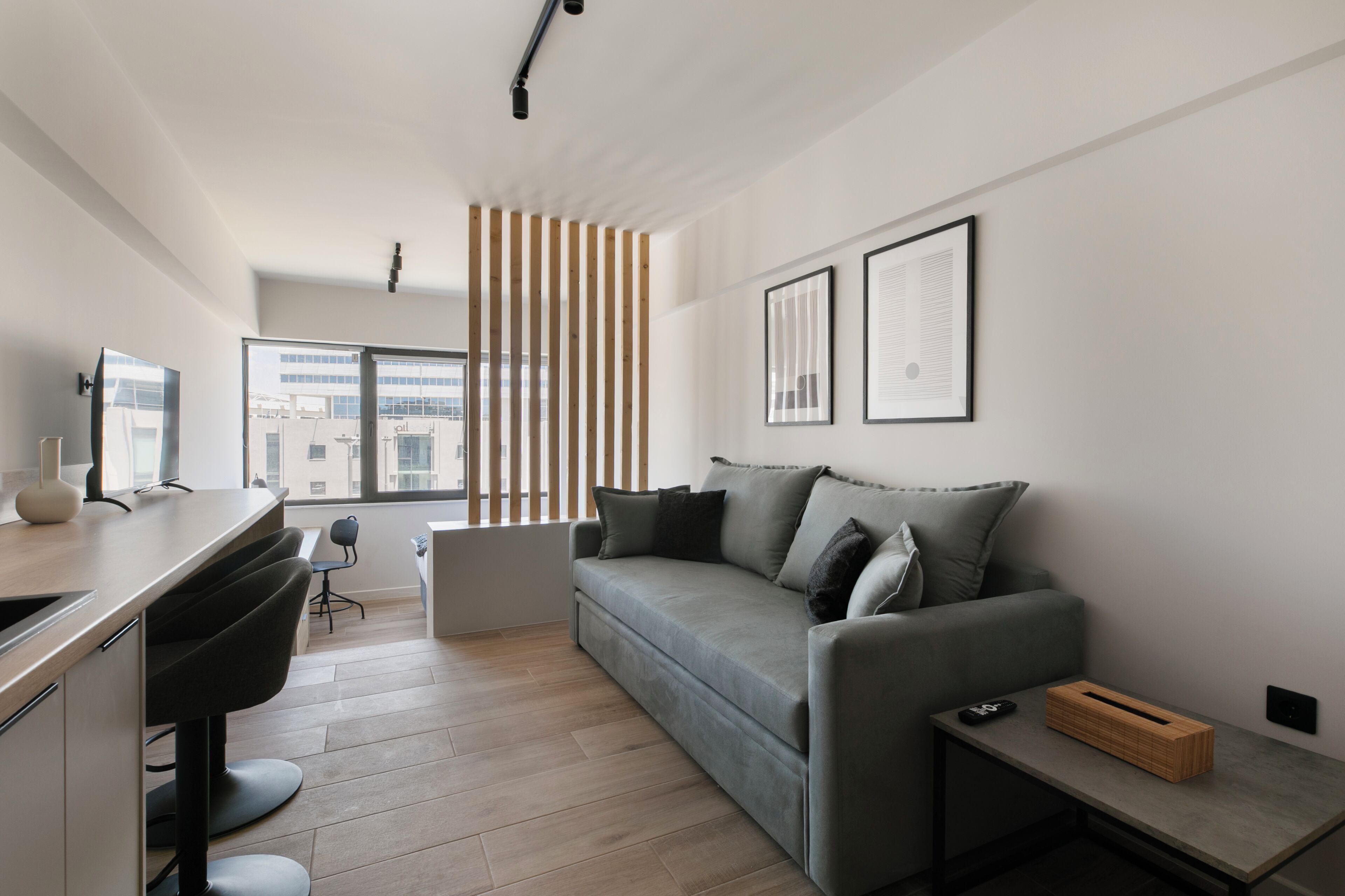 Ein modernes Wohnzimmer mit einem grauen Sofa, einer Trennwand aus Holzleisten, einem Schreibtisch mit Stuhl und einem kleinen Tisch mit Fernbedienung.