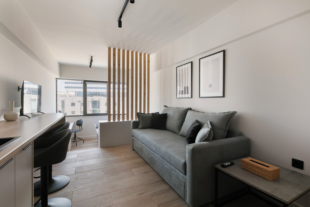 Ein modernes Wohnzimmer mit einem grauen Sofa, einer Trennwand aus Holzleisten, einem Schreibtisch mit Stuhl und einem kleinen Tisch mit Fernbedienung.