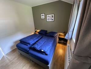 Een slaapkamer met een bed, twee nachtkastjes, een raam met gordijnen en twee ingelijste foto’s aan de muur.