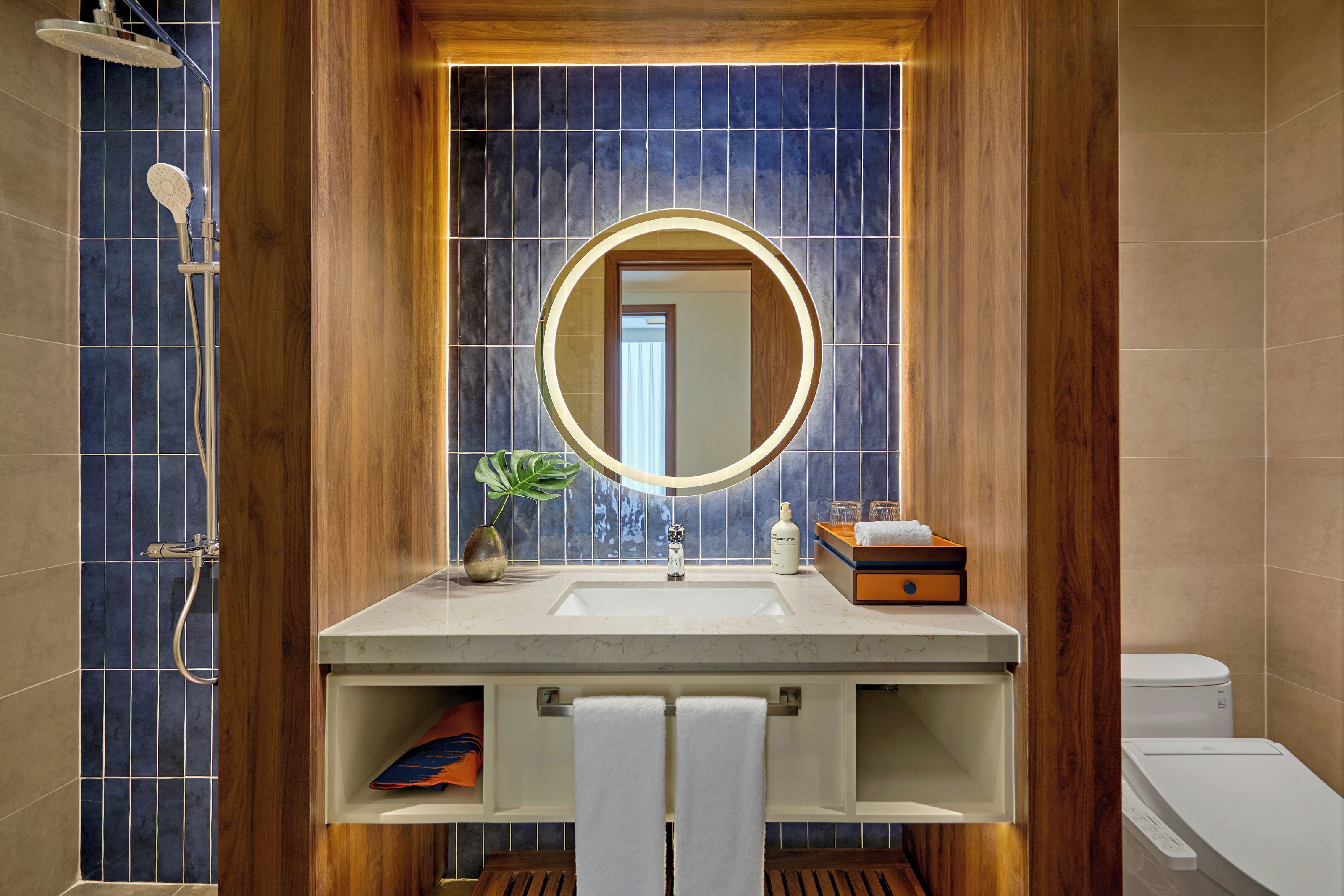 A bathroom with a wooden vanity, a round mirror, a shower, and a toilet.