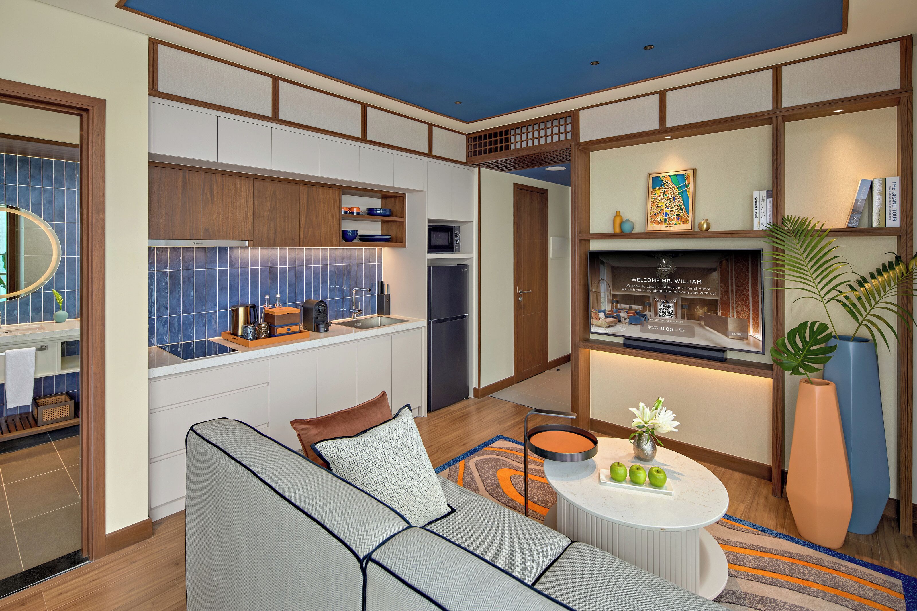 Una cocina moderna con microondas, horno y lavamanos, un área de estar con sofá y mesa de centro, y una estantería con libros y objetos decorativos.