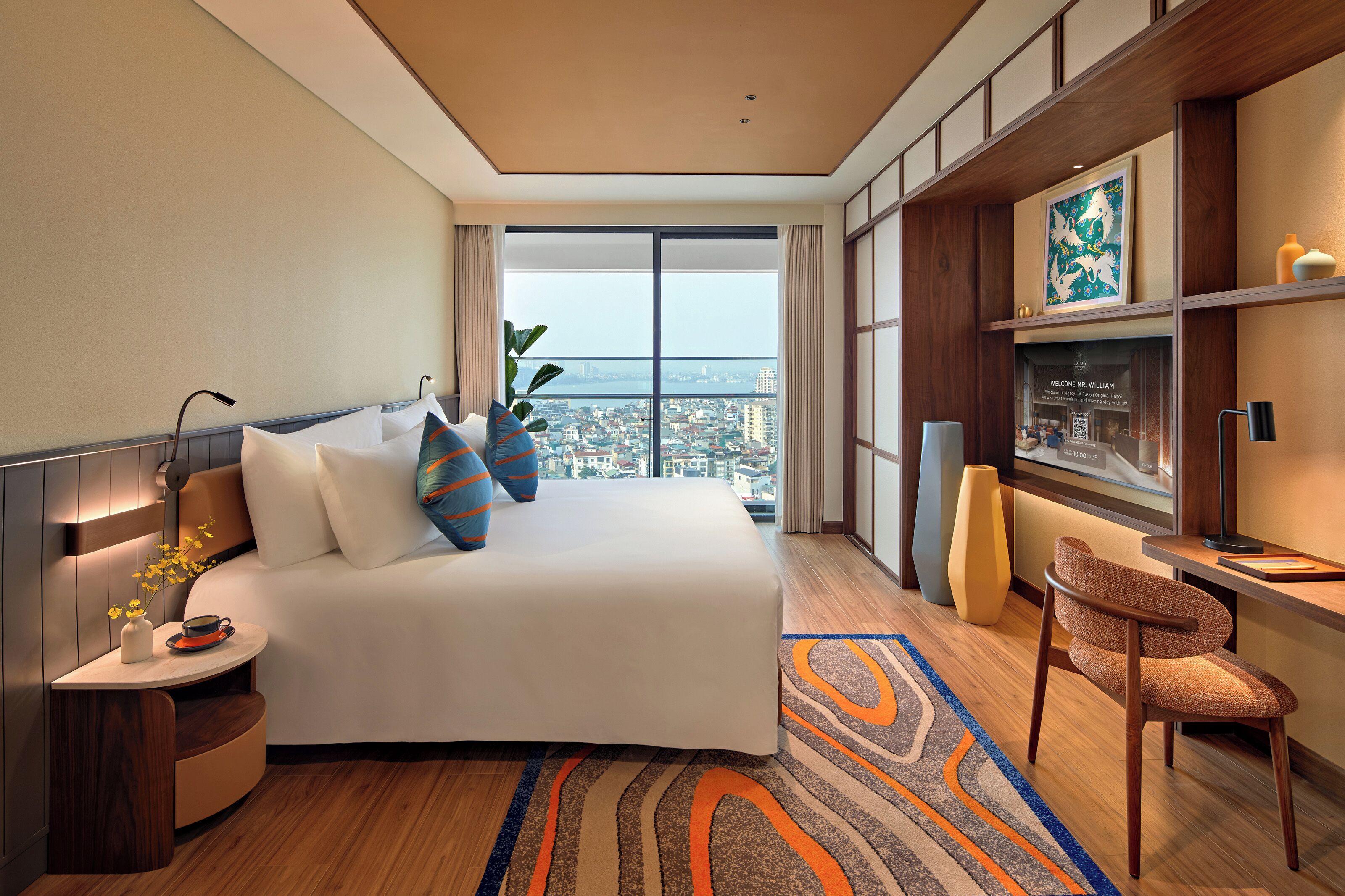 Quarto de hotel com uma cama grande, uma escrivaninha, uma cadeira, uma janela ampla com vista para a cidade e um quadro decorativo na parede.