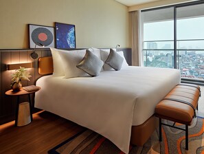 Quarto de hotel com uma cama grande, um banco, uma mesinha de cabeceira com abajur, um vaso com flores e vista para a cidade.
