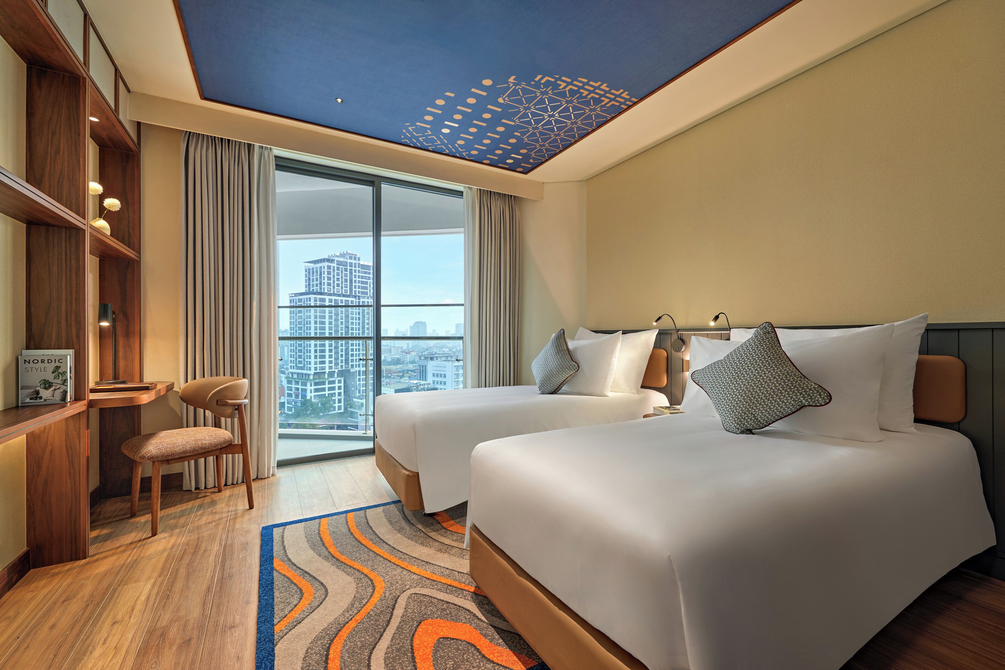 Quarto de hotel com duas camas, uma escrivaninha, uma cadeira, uma janela grande e vista para a cidade.