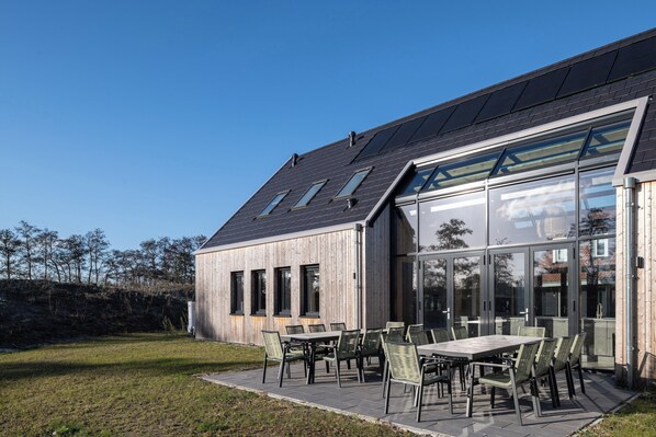 A modern building with a sloped roof, large windows, and a patio area with outdoor seating. - Ten-bedroom Group Villa Near the Shores (Nieuwvliet)