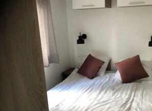 Ein Schlafzimmer mit einem Bett, zwei Kissen, einem Nachttisch, einer Lampe und einem Fenster mit Vorhängen.