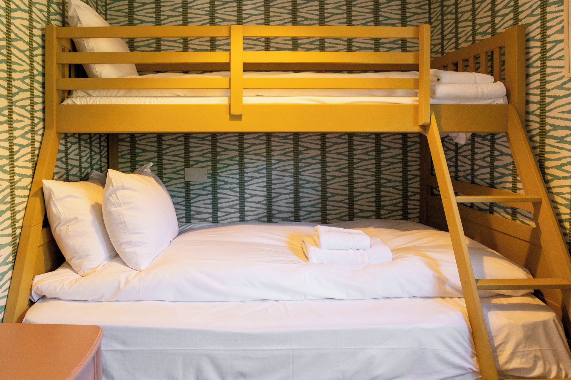 A bunk bed with white bedding and pillows, a wooden ladder, and a patterned wall.