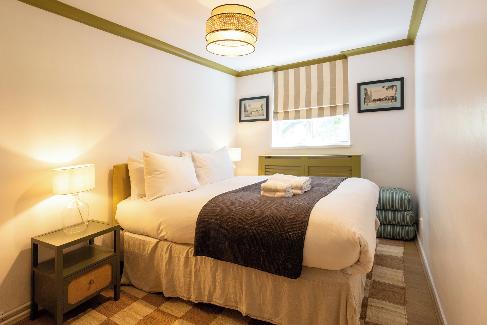 Habitación de hotel con cama, mesita de noche, lámpara, ventana con persianas y una pequeña silla verde.