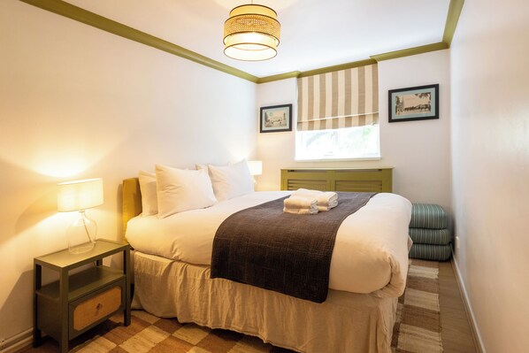 Una camera d'albergo con un letto, un comodino, una lampada, una finestra con tende e una piccola sedia verde.