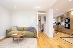 Eine moderne Wohnung mit einem Wohnzimmer, das ein Sofa, einen Couchtisch und gemusterte Kissen bietet. Ein Flur führt zu einer Küche mit Holzschränken und einem Herd.
