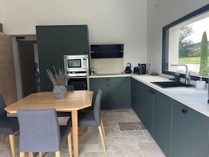 A modern kitchen with a built-in microwave, oven, and coffee maker, a dining area with a wooden table and chairs, and a window with a view of greenery. - A lovely house with a private pool, in a quiet location, 20 minutes from Lyon (Dardilly)