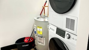 Un cuarto de lavado con lavadoras y secadoras apiladas, un calentador de agua y un cubo rojo.
