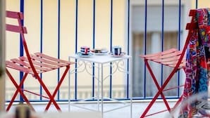 A balcony with red chairs and a small table, a railing, and a building in the background.