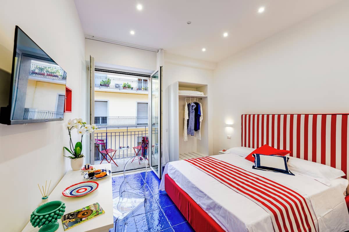 A hotel room with a bed, a TV, a balcony with a view of the building, and a dining area with chairs and a table.