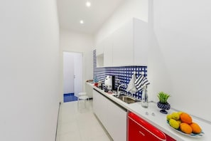 A narrow kitchen with white cabinets, a red dishwasher, and a blue tiled backsplash. - Bellamore (Piano di Sorrento)