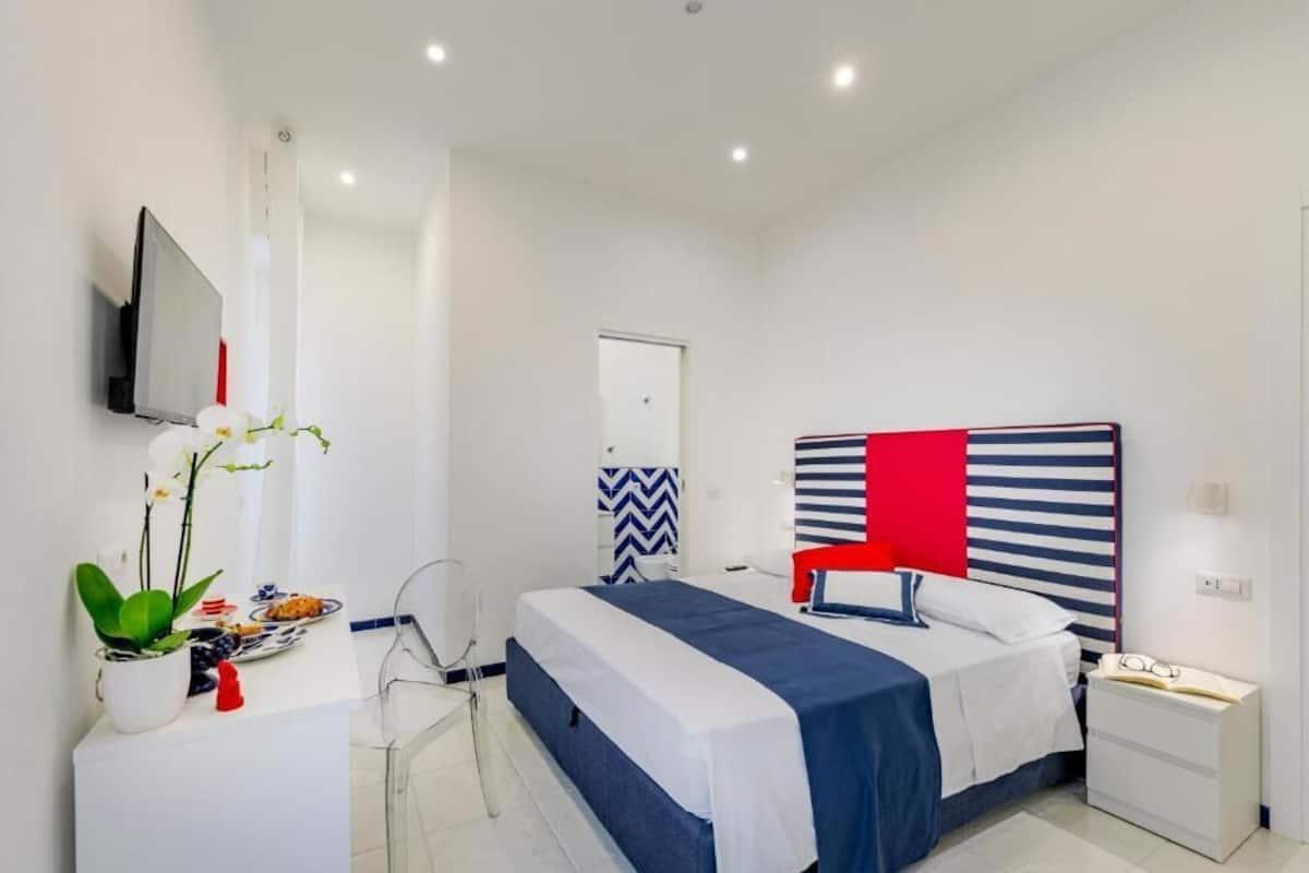 A modern hotel room with a bed, bedside table, chair, TV, and a decorative wall panel.