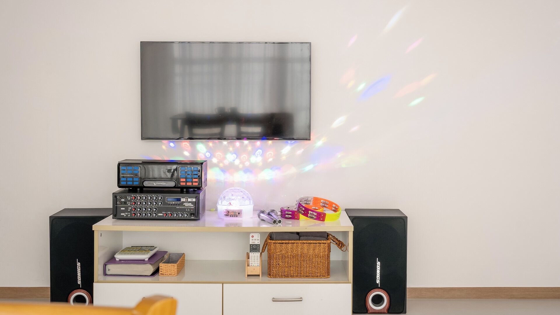Ett rum med en platt-TV som är monterad på väggen, en DJ-mixer, högtalare, en korg av rotting och ett litet bord med böcker och en fjärrkontroll.