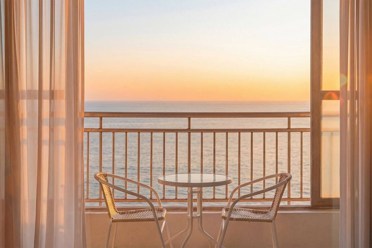 A balcony with two wicker chairs and a small round table, overlooking a body of water at sunset.