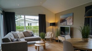 Ein modernes Wohnzimmer mit einer Couch, Sesseln, einem Esstisch, einem Fernseher und einem großen Fenster mit Vorhängen.