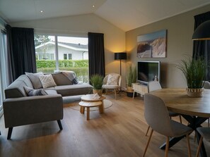 Ein modernes Wohnzimmer mit einer Couch, Sesseln, einem Esstisch, einem Fernseher und einem großen Fenster mit Vorhängen.