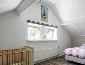 Ein Schlafzimmer mit schrägemausertem Dach, einem Fenster mit Aussicht, einem Babybett und einem Bett mit farbenfroher Tagesdecke.