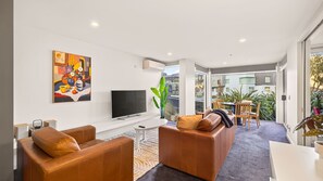 A modern living room with a flat-screen TV, leather sofas, a dining area with a table and chairs, and a large window offering a view of the city. - The Central Nook - Walk to town - Mountain views (Queenstown)