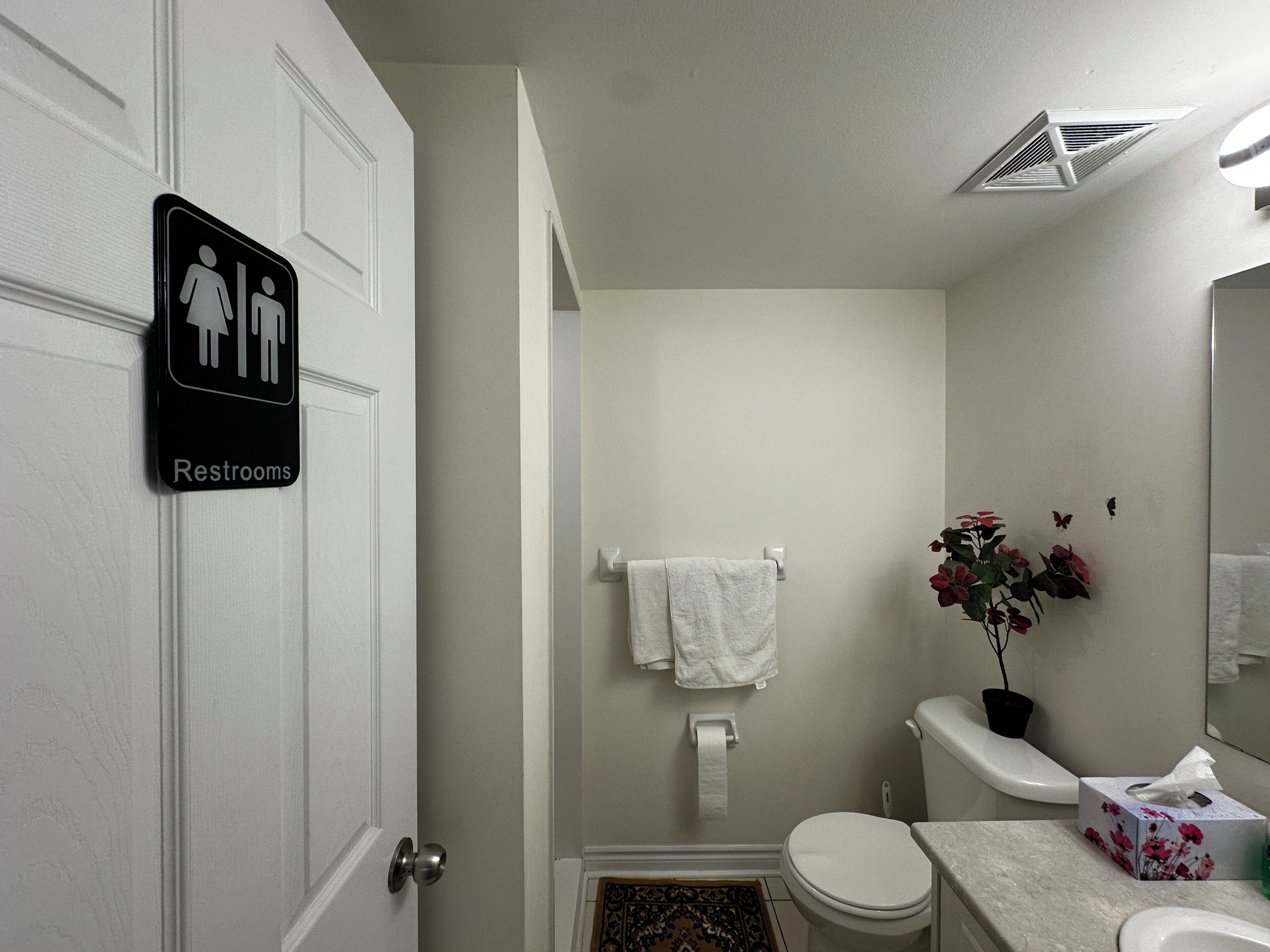 A bathroom with a toilet, a sink, a mirror, a towel rack, and a sign indicating restrooms.