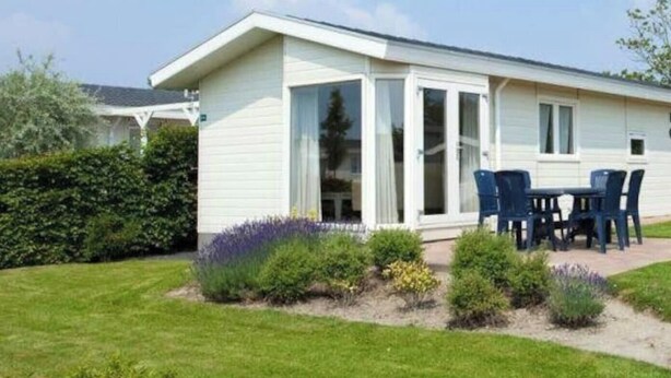 Ein eingeschossiges Mobilheim mit einer Terrasse, einem kleinen Garten mit verschiedenen Pflanzen und einem wolkenlosen Himmel.