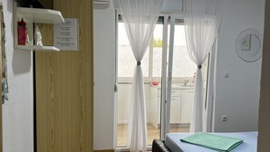 Quarto de hotel com uma cama, porta de madeira, janela com cortinas leves e vista para a varanda, além de um relógio na parede.
