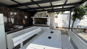 Uma cozinha ao ar livre coberta, com uma mesa comprida, bancos e uma lareira de pedra.