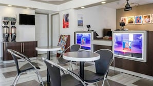 A modern hotel lobby with round tables, black chairs, a coffee machine, and a TV displaying a news channel.