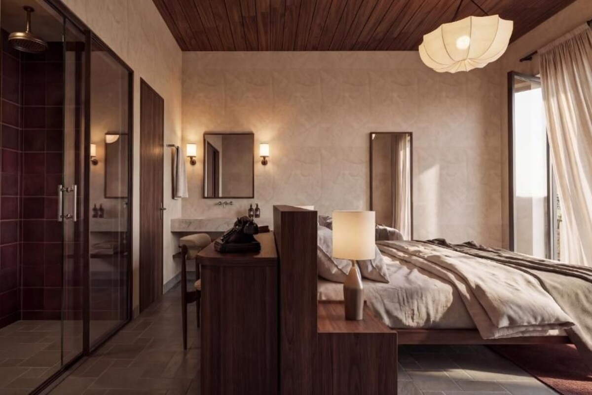 A modern hotel room with a shower, a bedside table, a lamp, a bed, and a large mirror.