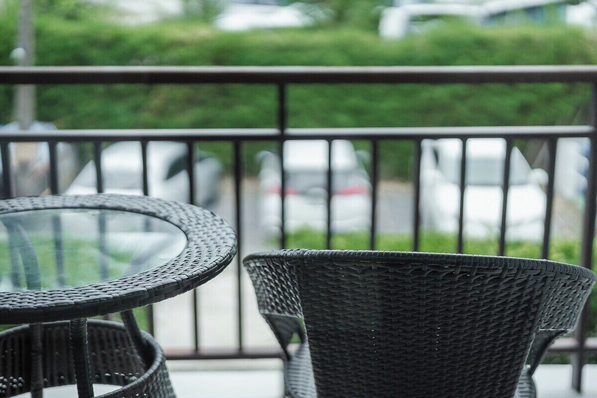 Un balcon doté d’une table ronde en verre et de chaises en osier, offrant une vue sur un parking et sur un espace verdoyant.