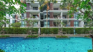 A swimming pool surrounded by a multi-story building with balconies, a stone wall, and greenery. - Beachside 1BR•Walk to Rawai•Cozy-title rawai C201 (ตำบล ราไวย์)