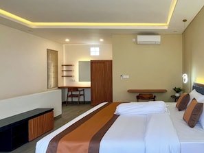 A hotel room with a bed, a desk, a chair, a small table, and a potted plant.